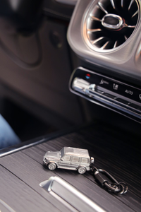 Schlüsselanhänger in Form eines silbernen Mercedes-Benz G-Modells, abgebildet auf einer stilvollen schwarzen Oberfläche im Fahrzeuginterieur. 1