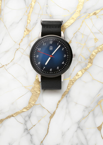Elegante Armbanduhr mit blauem Zifferblatt, Mercedes-Benz-Logo und rotem Sekundenzeiger, an einem schwarzen Textilarmband befestigt, präsentiert auf einer luxuriösen weißen Marmortextur mit goldenen Adern. 1