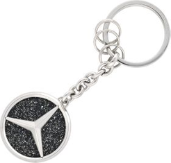 Schlüsselanhänger aus Metall mit dem Mercedes-Benz-Logo, auf einer glitzernden, schwarzen Oberfläche.